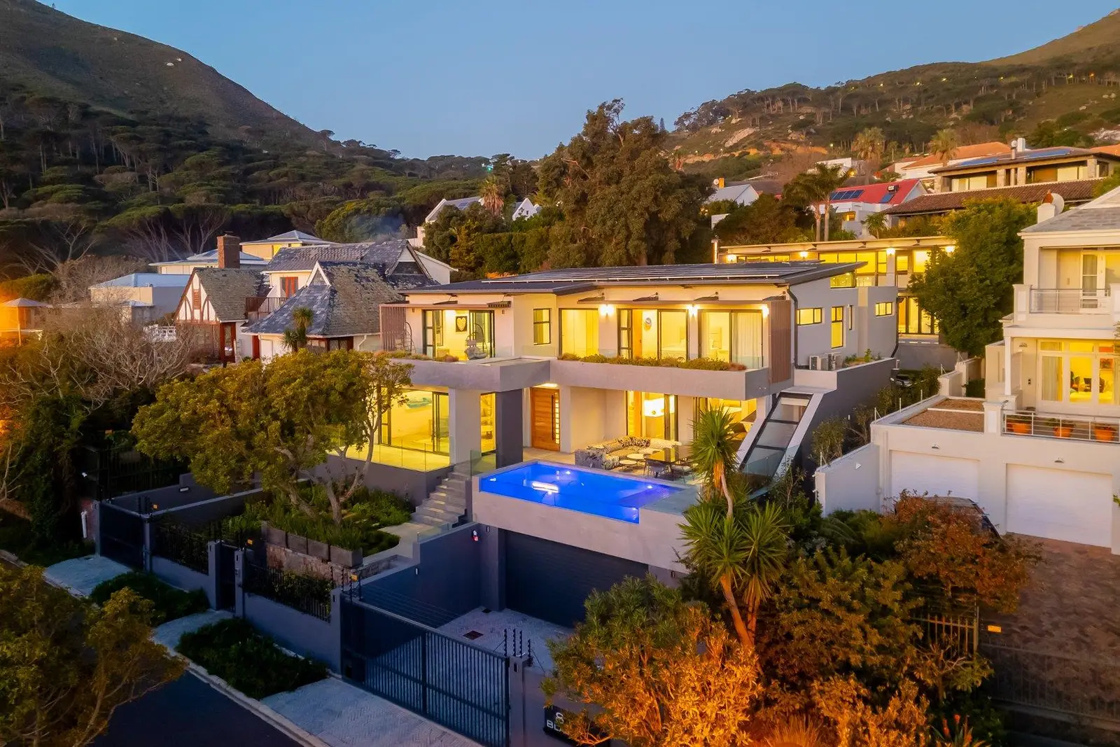 Large modern house with lit windows at dusk, a lit blue pool on the terrace and wooded mountain slopes behind