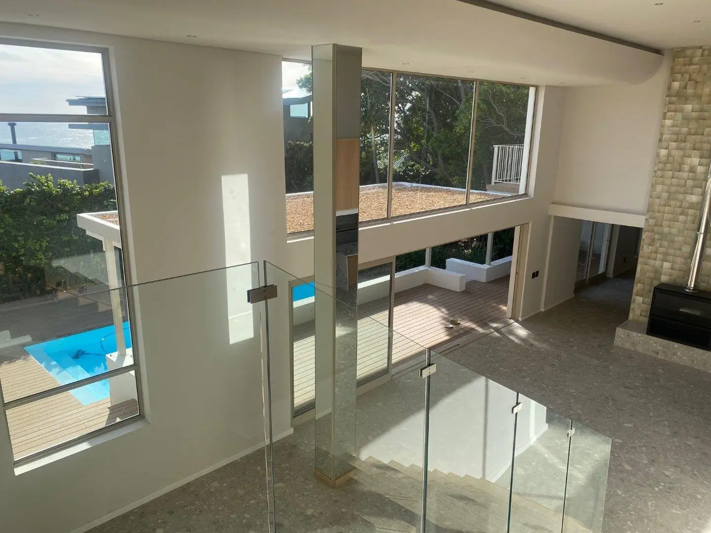 Double-height room seen from a landing behind a glass balustrade, with tall windows onto a timber deck and a blue pool, and a stone-clad fireplace on the right