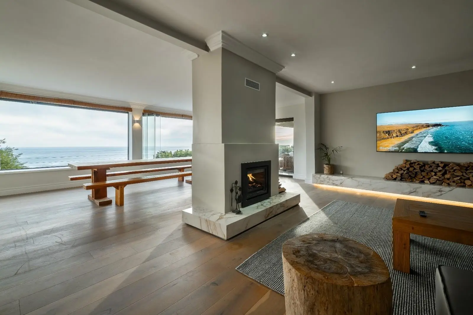 Open living room with a double-sided fireplace, a long wooden dining table and wide windows looking out to the sea